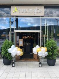 Eine zweiflügelige offenstehende Türe. Davor rechts und links Luftballons und zwei hohe Topfpflanzen.
Über dem Eingang hängt in großen Lettern "Hans im Glück" mit Firmenlogo.