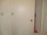  weiße einflügelige Türe in einer weißen Wand, rechts davon ist eine schmale Glasscheibe. Links davon in Kopfhöhe ein Türschild
