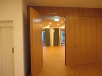offenstehende Holztür in einer Holzwand mit Blick in den Gemeindesaal. Links im Vordergrund ist ein Teil der Aufzugstür