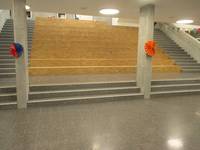 breite Sitzstufen aus hellem Holz, über 3 Stufen erreichbar.  an den Stufen sind 2 Betonpfeiler, links und rechts von den Sitzstufen sind Treppen. In der Decke sind runde Lichter