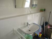 Ein weißes Waschbecken an einer weiß gekachelten Wand. Links ein Haltegriff und rechts auf einer Stange ein Handtuch. Über dem Waschbecken ein kippbarer Spiegel.