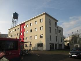 vierstöckiges Flachdachgebäude, auf Längsseite ist in der Mitte ein zurückgesetzter, rot gestrichener Teil. Vor dem Gebäudes ist ein Parkplatz und ein Fahrradständer, hinter dem Haus befindet sich ein Wasserturm
