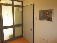 helle Tür mit dunklem Rahmen, Türklinke niedrig angebracht. Rechts neben Tür helle Wand mit Türschild und Bild, links von der Tür Milchglasfront