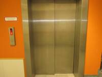 Geschlossene Aufzugtüren aus Edelstahl in einer orangefarbenen Wand. Links von Aufzug Anforderungstaste mit Anzeige des Stockwerks E. Darüber hängt ein Schild mit der Aufschrift: Aufzug im Brandfall nicht benutzen