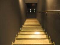 Eine abwärts führende Treppe mit hellen Stufen zwischen zwei schwarzen Wänden. Rechts ist ein Handlauf.