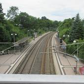 zwei Schienenstränge, mit Bahnsteigen rechts und links, auf den Bahnsteigen überdachte Wartebereiche, hinter den Bahnsteigen sind auf beiden Seiten Bäume