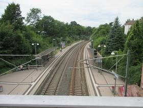 zwei Schienenstränge, mit Bahnsteigen rechts und links, auf den Bahnsteigen überdachte Wartebereiche, hinter den Bahnsteigen sind auf beiden Seiten Bäume