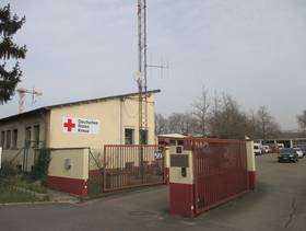 Ein- und Ausfahrt getrennt durch ein Metalltor, links ein Gebäude mit einem angeschrägten Flachdach mit dem Roten Kreuzzeichen an der Seitenwand, dahinter der Hof mit Parkplatz des Roten Kreuz