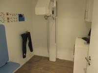 kleiner dunkler Raum mit höhenverstellbarem Röntgengerät an hinteren Wand, links davon an der Wand befestigter Behandlungsstuhl 