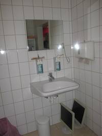 Waschbecken mit Seifenspender und Desinfektionsmittel an der Wand, darüber hängt ein rechteckiger rahmenloser Spiegel 