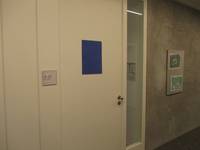 weiße Tür, rechts daneben schmale bodentiefe Glasscheibe. Graue Wand mit einem Bild in einem Glasrahmen, auf der Tür ist ein blauer DIN A4 Aushang