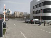 Ein großer Parkplatz mit zwei Schranken. Rechts ist ein mehrstöckiges Gebäude mit silberfarbener Fassade
