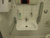 Waschbecken mit Handlauf rechts und links, darüber ein kippbarer Spiegel mit einem Zugseil. Links an der Wand ein Papiertuchhalter. Über dem Waschbecken ist ein Seifenspender