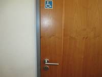 Holztür mit Drücker links, Aufschlag nach außen. Links oben blaues Schild mit Rollstuhl-Symbol