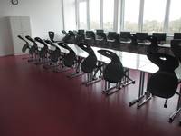 doppelte Tischreihe mit aufgestellten Stühlen, hinten an der Wand eine breite Fensterfront mit weiteren Tischen, auf denen Bildschirme stehen