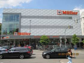 großes 3-stöckiges Kaufhausgebäude mit Flachdach, EG mit durchgängiger Schaufensterfront und Eingangsbereich, ab 1. OG weiße Kunstoffverkleidung, Linke Gebäudeecke im 1. und 2. OG verglast. Schriftzug "Müller" mit Logo rechts an Dach und links übe