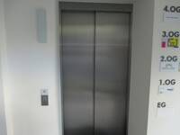 Ein geschlossener Aufzug mit Edelstahltür, rechts daneben Schilder mit Hinweisen zu Einrichtungen in den verschiedenen Etagen