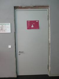 Tür zur Behindertentoilette im Flur 2. Die Tür und der Türrahmen sind weiß. Auf der Tür-Mitte ist das Toilettenschild aufgeklebt. Direkt neben der Toilettentür hängt an der Wand ein Flucht- und Rettungsplan. 
