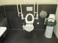 Eine weiße Toilette an einer schwarzen Wand, auf jeder Seite ist ein hochgeklappter Haltegriff. Rechts neben der Toilette steht ein großer Hygienbehälter.