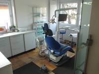 Raum mit verschieden Regalen an den Wänden, in der Mitte steht ein Zahnarztstuhl mit einer blauen Sitzfläche. Auf dem Boden liegen Schmutzfangmatten