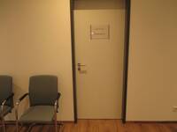 Eine helle Türe in einem dunklem Rahmen in einer hellen Wand. Auf der Tür ist ein Schild mit der Aufschrift: Arztzimmer 3.  Links vor der Tür stehen 2 Stühle an der Wand.