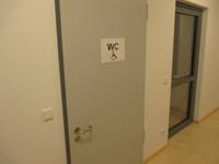 hellgraue Türe mit dunklem Rahmen in weißer Wand, auf der Tür ist ein Schild mit Rollstuhlsymbol und dem Schriftzug WC, rechts davon ist eine Brandschutztür