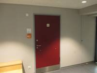 Rote Tür vor hellen Wand, auf der Tür ist ein Rollstuhlfahrersymbol, links an der Wand ein Feuermelder und ein Schild