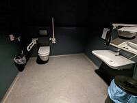 Hängetoilette, mit Haltegriff rechts und links, an der Wand Toilettenpapier und Klobürste und Mülleimer, rechts Waschbecken mit Spiegel, Seifenspender links und Mülleimer rechts