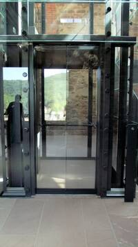 komplett verglaste Kabine, Tür öffnet mittig. Rechts neben dem Aufzug schwarze Stele mit Anforderungsknopf