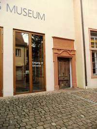 Im Vordergrund ist ein Platz mit Kopfsteinpflaster zu sehen daran anschließend ein Ausschnitt des Museumgebäude, mit einem großen Fenster und der zweiflügeligen Eingangstür zum Museum