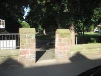 Halbhoge Mauer mit zwei Steinpfeilern rechts und links von einem offenen Durchgang. Dahinter der Park mit Bäumen und Wegen
