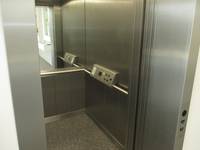 Offener Aufzug mit Metallwänden und Steinboden, rechts ist ein Handlauf mit einer Tastatur. Der Handlauf setzt sich fort und an der Rückwand ist ein Spiegel
