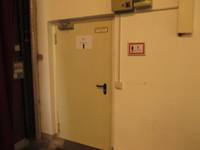 weiße einflügelige Tür in einer weißen Wand: Auf der Tür ist ein DIN-A4 Aushang, rechts neben der Tür ist ein Schild mit einem roten Rand und ein Kabel, das zu einem Lichtschalter führt