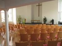 Großer Kirchenraum mit Altar und schlcihten Kreuz, davor und an beiden Seiten dichte Bestuhlung mit Holzstühlen in mehreren Reihen 