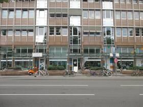 mehrstöckiges Gebäude, im Erdgeschoss Schaufensterfront, davor Pflanzenkübel, Fahrräder und die Straße