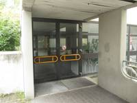 zweiflügelige Glastür mit gelben Querstangen, der Bereich davor ist überdacht. Vor der Tür Fußabstreifer aus Metallgitter