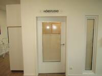 Eine Glastür mit einem weißen Rahmen in einer weißen Wand. Über der Tür ist ein Schild aus Plexiglas mit der Aufschrift: Hörstudio.
