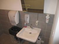 Waschbecken mit Spiegel, links Mülleimer und Papierhandtücher, über dem Waschbecken linksseitig ein Seifenspender 