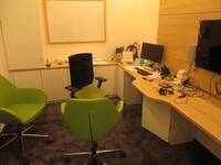 Ein Raum mit einem längeren Schreibtisch an der rechten Wand, davor stehen 3 grüne Stühle