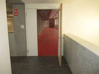 offenstehende Tür in einem weißen Rahmen, rechts neben der Tür ist eine halbhohe Mauer. Links geht es in einen Flur, hinter der Tür ist der rote Boden des Gymnastikraums
