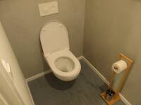 Eine weiße Toilette an einer hellgrauen Wand. Vor der Toilette ist ein hoher Toilettenpapierhalter aus Holz.