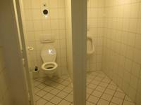 Zwei offen stehende schmale Toilettenkabinen. In der linken Kabine ist eine Hängetoilette, in der rechten ein Pissoir 
