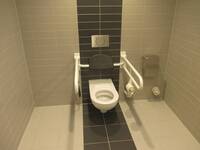 Eine weiße Toilette mit je einem Haltegriff auf jeder Seite. Der Raum ist hell gefliest, bei der Toilette ist auf dem Boden und bei der Wand ein schwarz gefliester Streifen.