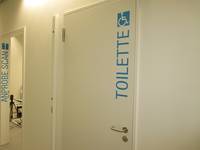 Weiße Tür in einer weißen Wand. Oberhalb der Türklinke steht in großer blauer von unten nach oben senkrecht angeordneter Schrift: Toilette. Darüber ein Rollstuhlsymbol