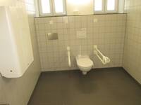 Eine weiße Toilette an einer weiß gekachelten Wand. Auf beiden Seiten ist je ein Haltegriff. Links ist ein klappbarer Wickeltisch.