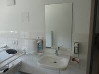 Weißes Waschbecken an weißer Wand. Darüber hängt ein Spiegel. Links vom Waschbecken Behindertentoilette mit Haltegriffen.