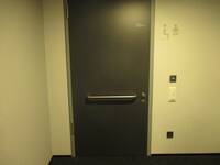 Eine dunkle Tür mit einer waagrechten Griffstange in einer hellen Wand. Rechts von der Tür ist eine weiße TAste zum automatischen Öffnen der Tür.