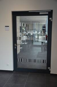 breite Glastür mit dunklem Metallrahmen in weißer Wand. auf der Tür sind konstrastierende Bereichen auf 2 Höhen angebracht. der obere Streife ist durch die Aufschrift Business-club" unterbrochen