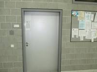 helle einflügelige Tür in einer grauen Wand, rechts eine Pinwand mit Aushängen