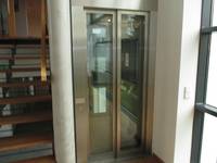Aufzug mit Glaskabine, Tür geschlossen. Links neben Aufzug aufwärts führende Treppe mit offenen Holzstufen